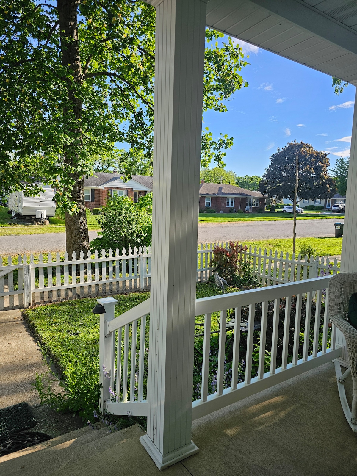 A view from a covered porch features a white picket fence and a well-maintained lawn. A large tree provides shade to the yard, while residential homes are visible in the background. The sky is clear, revealing bright sunlight.