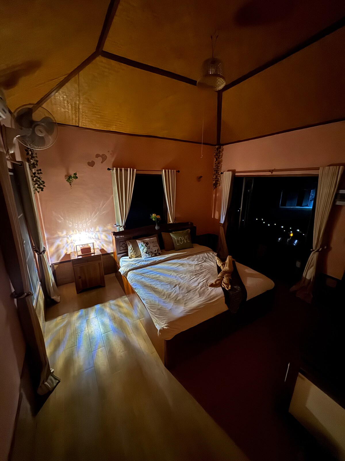 A spacious sleeping area features warm, pink walls and wooden furnishings. Large windows allow soft light to enter, highlighting the neatly made bed with relaxed linens. A cozy ambiance is created by the subtle glow from a bedside lamp, enhancing the tranquil setting.