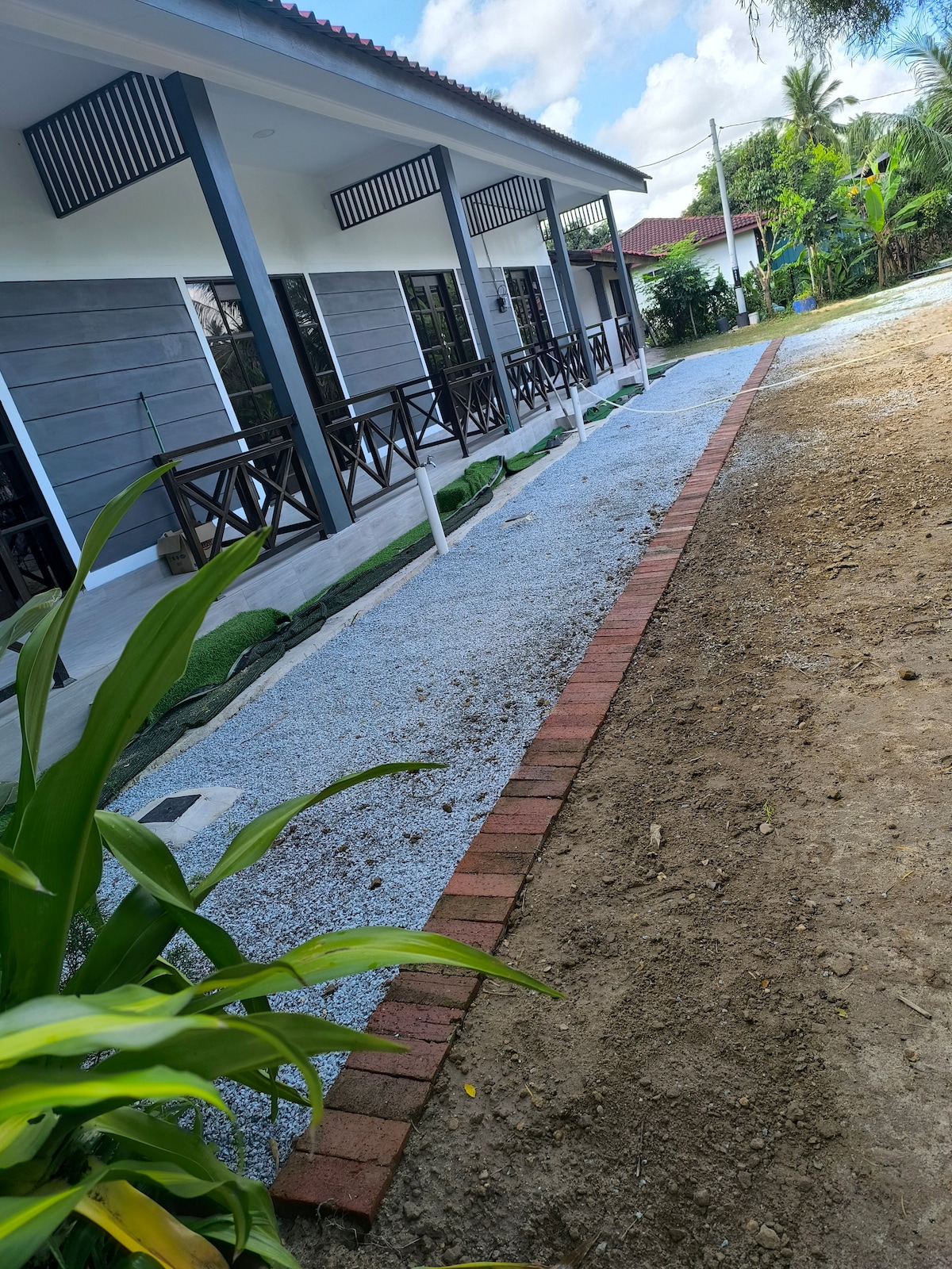 A garden path lined with bricks is featured, leading towards a modern chalet structure. The chalet has multiple doors and balconies, framed with dark railing. Lush greenery surrounds the area, enhancing the outdoor space's natural feel.