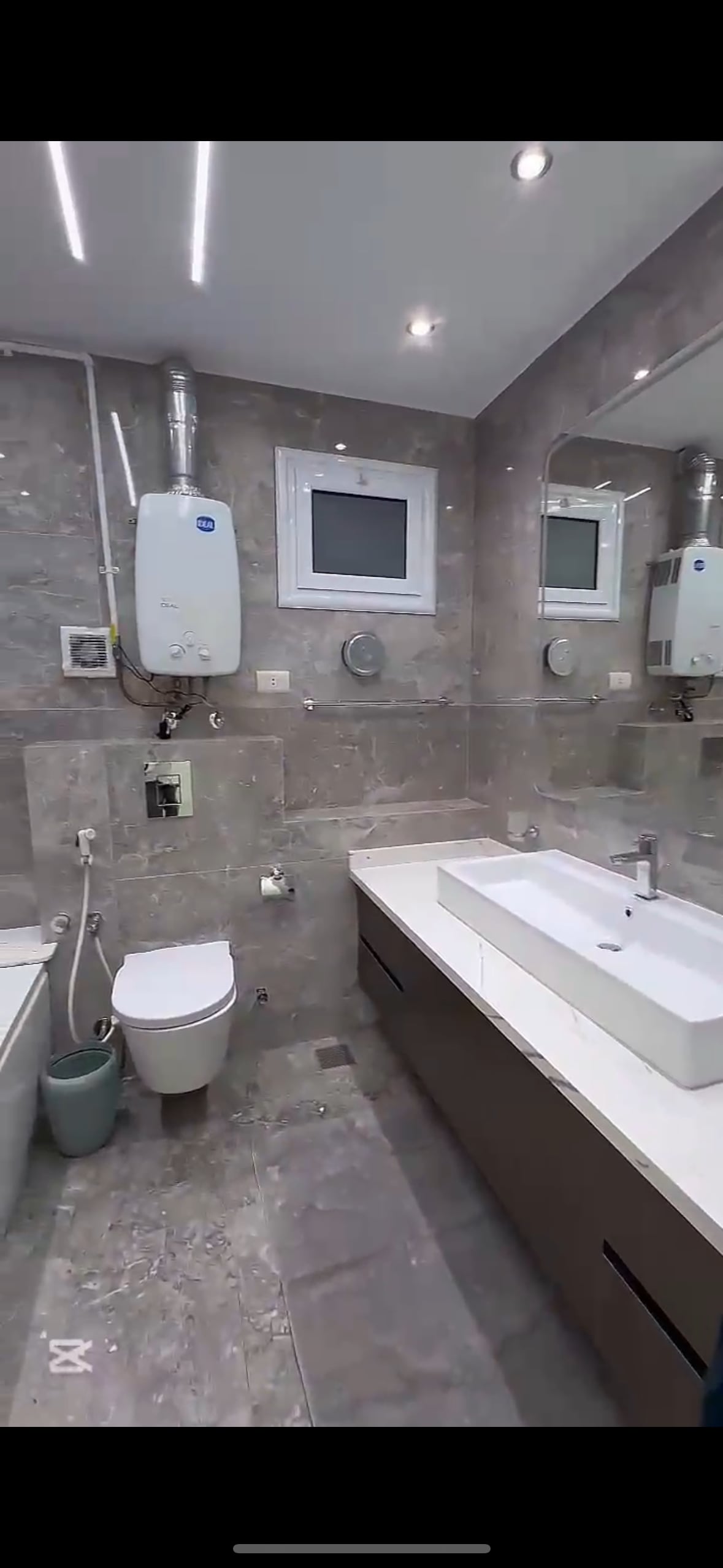 A modern bathroom features sleek grey tiles with a contemporary design. A spacious vanity with a wide sink is situated against the wall, while a mirror hangs above it. Bright lighting illuminates the space, complemented by minimalistic fixtures, including a toilet and storage details.
