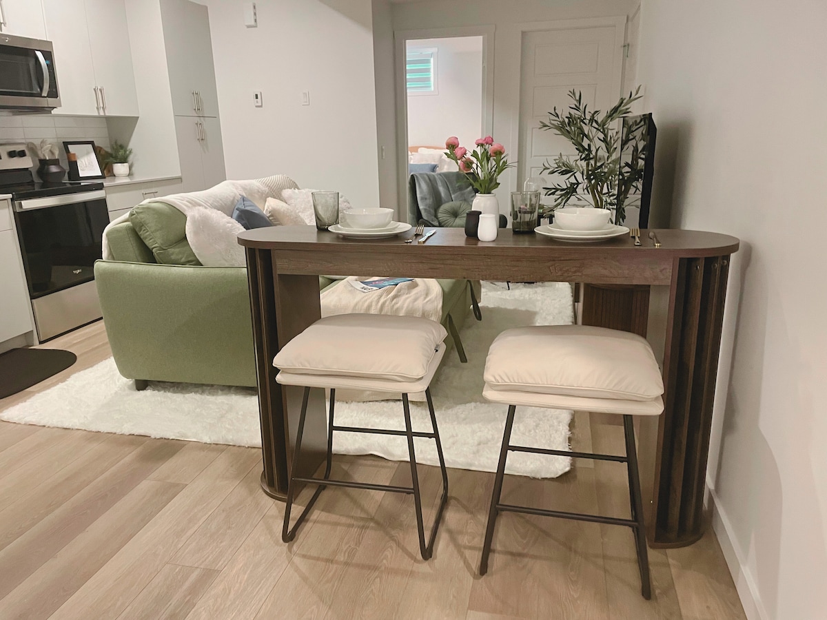 A modern dining area is composed of a stylish bar-height table with two cushioned stools. In the background, a cozy living area with a green sofa is visible, accompanied by a light neutral rug and a decorative arrangement of flowers on the table.