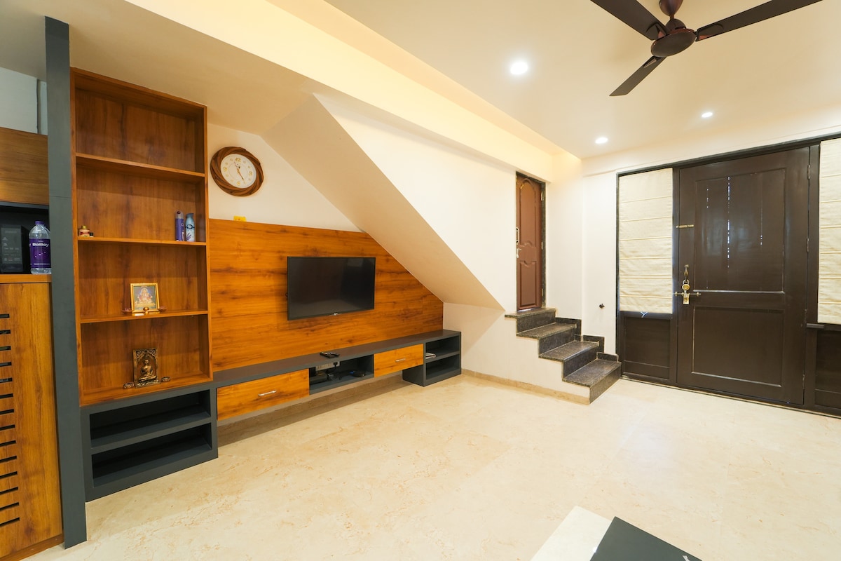 An open living space is showcased with a wooden accent wall, featuring a mounted television and decorative shelving. Natural light fills the room through a front door with glass panels, while a staircase leads to a higher level. A clock hangs above the shelves.