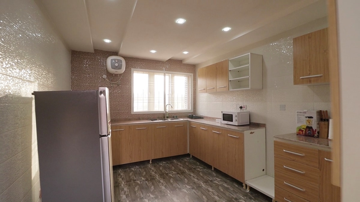 The kitchen features wooden cabinets and modern appliances, including a refrigerator and a microwave. A sink is positioned beneath a window, allowing natural light to illuminate the space. The backsplash reflects light, enhancing the overall brightness of the area.