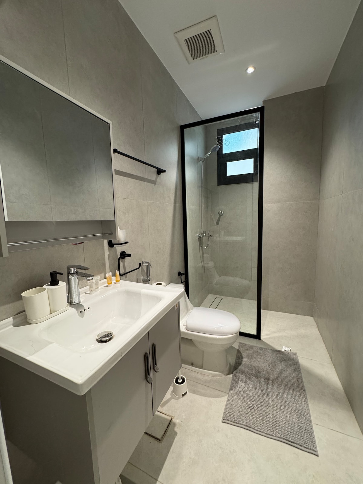 A modern bathroom features sleek gray wall tiles and a glass-enclosed shower. A white sink with silver faucets is positioned beneath a mirror, while a white toilet is located beside it. A light gray rug is placed in front of the sink area.