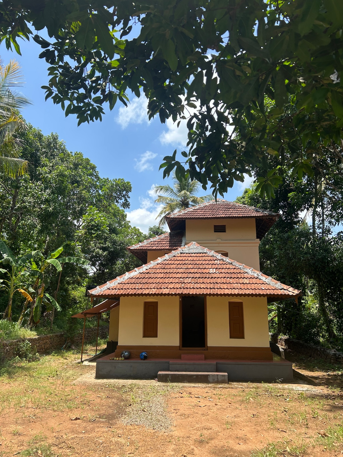 A traditional Kerala home is showcased among lush greenery, featuring a sloped terracotta roof. The exterior walls are painted in a soft hue, with multiple wooden windows. The surroundings consist of tall trees and tropical plants, enhancing the tranquil atmosphere of the property.