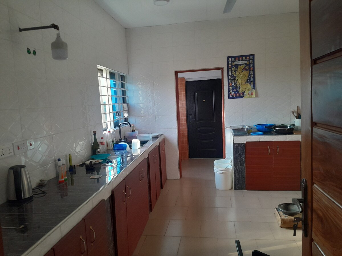 A spacious kitchen features sleek countertops and wooden cabinets. Natural light enters through multiple windows, illuminating the area. Household appliances, including a kettle and kitchenware, are neatly arranged, while a door leads to another room, creating an open and functional space.