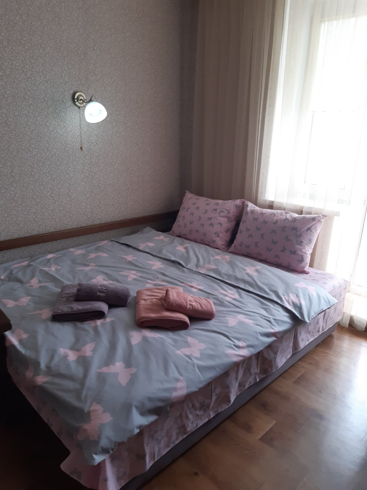 A bright sleeping area features a spacious bed dressed in butterfly-patterned bedding. Two decorative pillows complement the blanket, which is arranged casually. Light filters through a nearby window, illuminating the wooden floor and creating a soft ambiance.