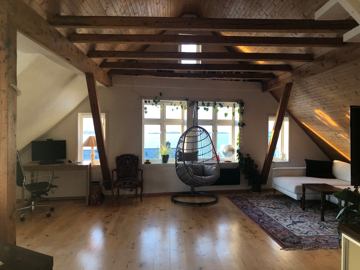 An airy attic space features wooden beams and a light wooden floor. A suspended hanging chair occupies the center, surrounded by large windows allowing ample natural light. A desk and lamp are positioned to one side, with a white couch and decorative plants completing the inviting atmosphere.