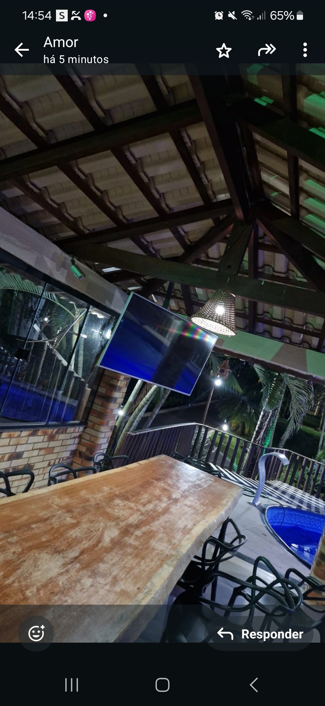 A wooden dining table is positioned under a sloped ceiling, surrounded by several black chairs. A large television is mounted on the wall, with surrounding windows offering views of the outdoors. A pool is visible in the background, enhancing the space's recreational potential.