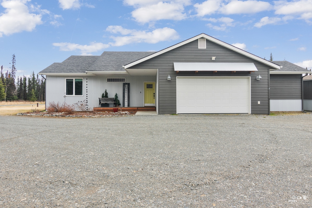 The exterior view of a modern, single-story home is presented, featuring a light gray exterior with a contrasting dark roof. A spacious driveway leads to a two-car garage, while a small porch is adorned with minimal landscaping and a welcoming yellow front door.
