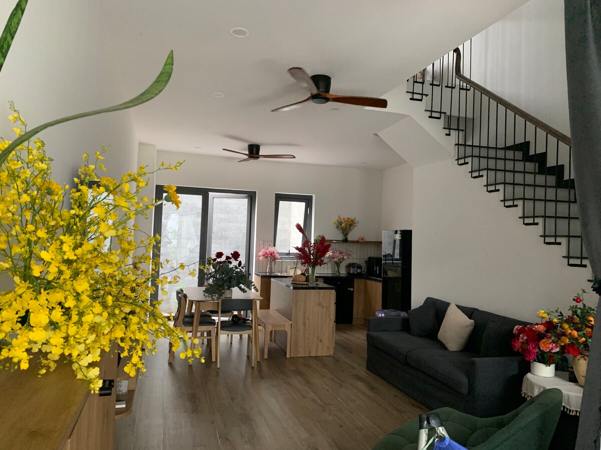 An open living area is highlighted by stylish ceiling fans and large windows that bring in natural light. A dark sofa is positioned beside wooden furniture, while vibrant floral arrangements in various colors add a touch of brightness to the space.