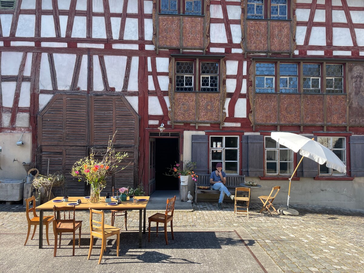 An outdoor seating area features a large wooden table surrounded by several chairs. A parasol provides shade, while vibrant flowers in vases decorate the table. A person is seen sitting casuallyNearby, the historic building showcases exposed timber beams and colorful window panes.
