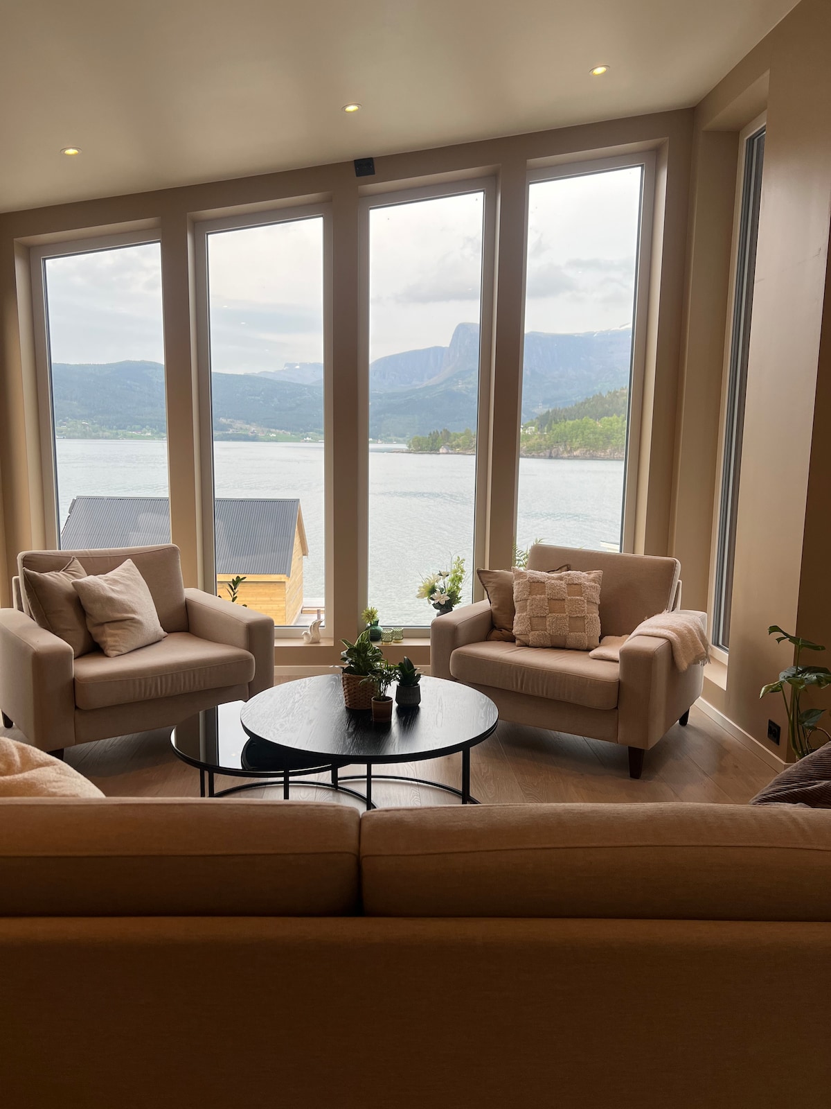 A comfortable seating area is framed by large windows overlooking the water and mountains. Two light-colored armchairs are positioned around a circular black coffee table, creating an inviting space. Natural light floods the room, enhancing the neutral color palette and serene views.