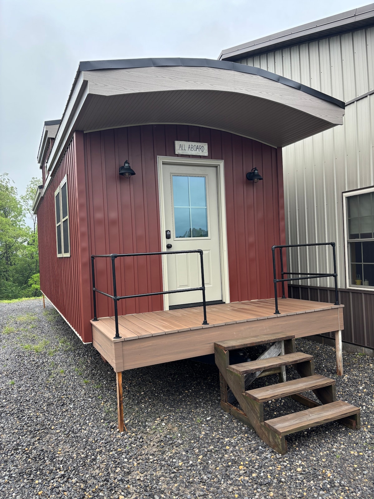 The exterior of the tiny house features a red metal facade with a welcoming porch area. A simple set of stairs leads to the entrance, which is adorned with a light fixture and a sign above the door. Surrounding greenery is visible in the background.