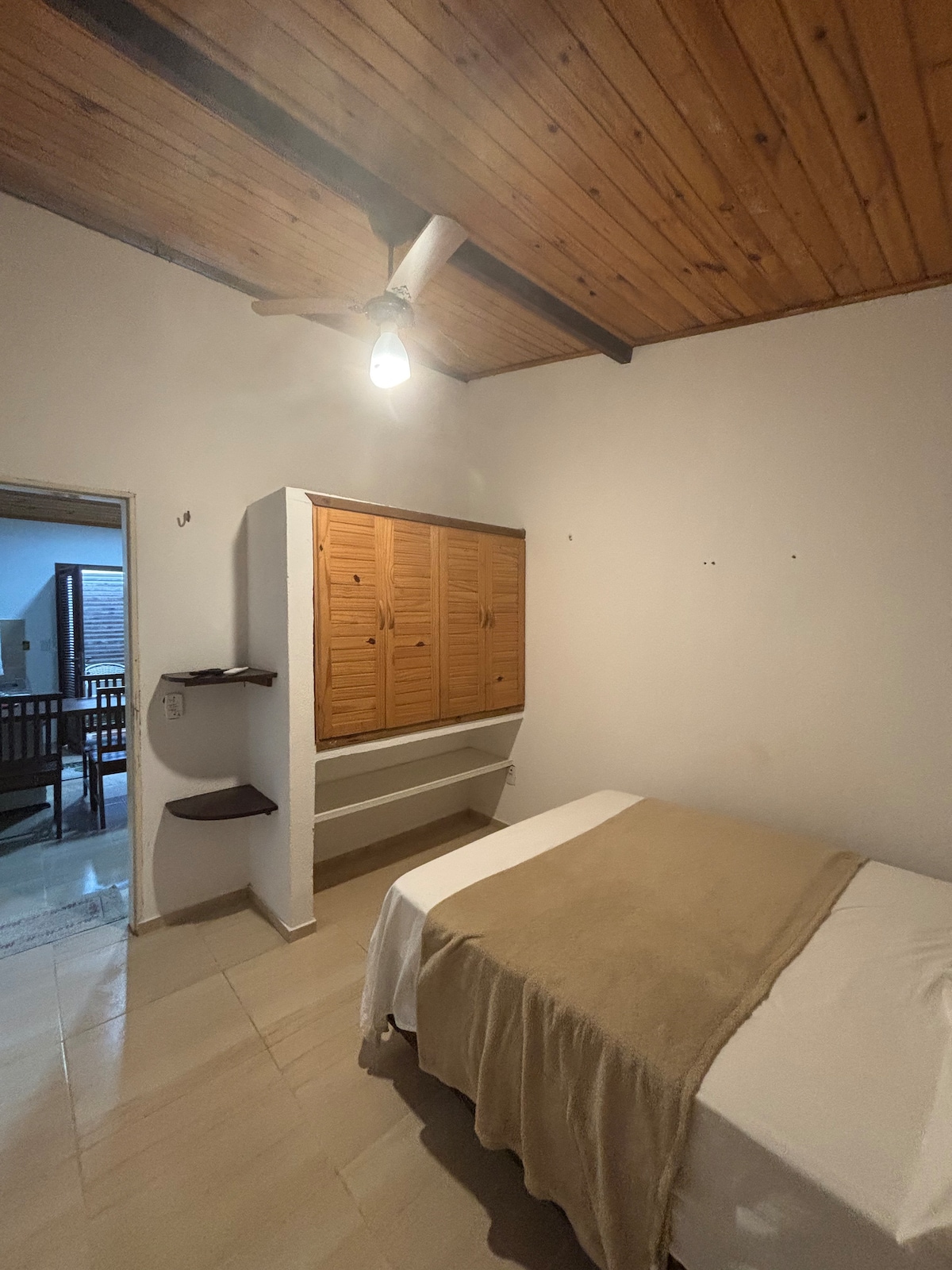 A well-lit bedroom features a spacious bed covered with a neutral-colored throw. Wooden cabinets line one wall, with a small shelf for additional storage. The ceiling displays wooden beams, adding warmth to the room, which connects to a communal living area.