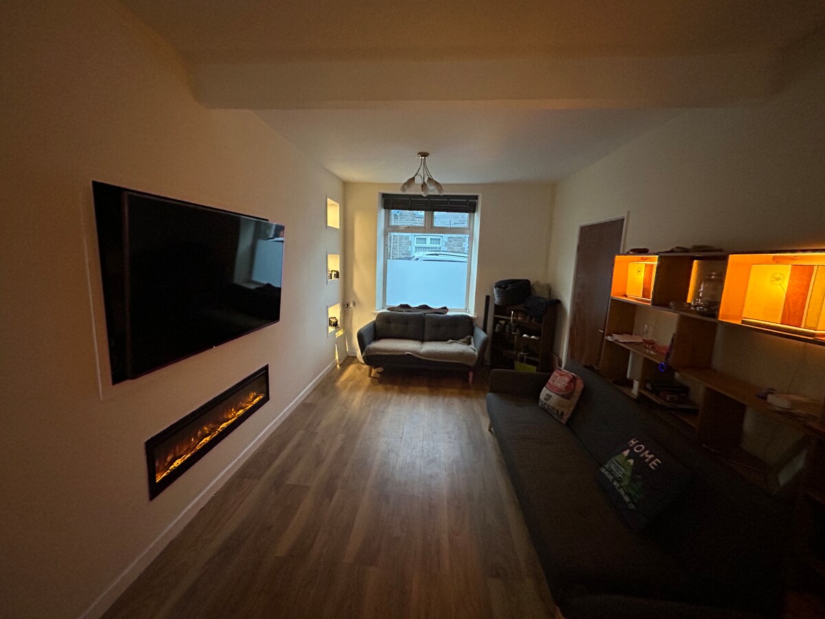 A modern living area features two sofas positioned around a wall-mounted television. A sleek electric fireplace is installed along the wall, providing a contemporary touch. Soft lighting enhances the calm atmosphere, while a window allows natural light to filter into the room.