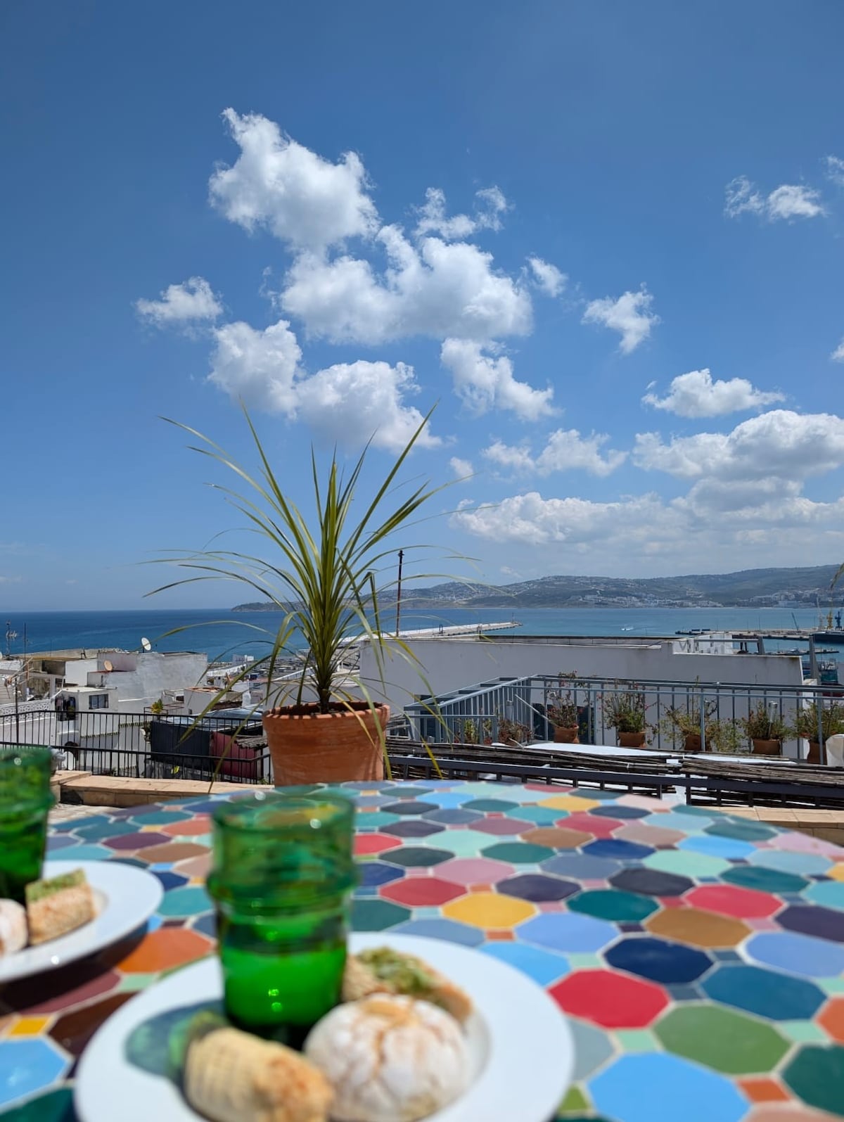 A colorful tiled table is visible in the foreground, adorned with green glasses and a small plate. Behind, a potted plant adds a touch of greenery. The background features a panoramic view of the sea, distant hills, and a partly cloudy sky.