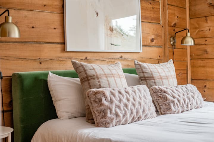 Acogedor dormitorio de cabaña con ropa de cama lujosa, detalles a cuadros y apliques de latón