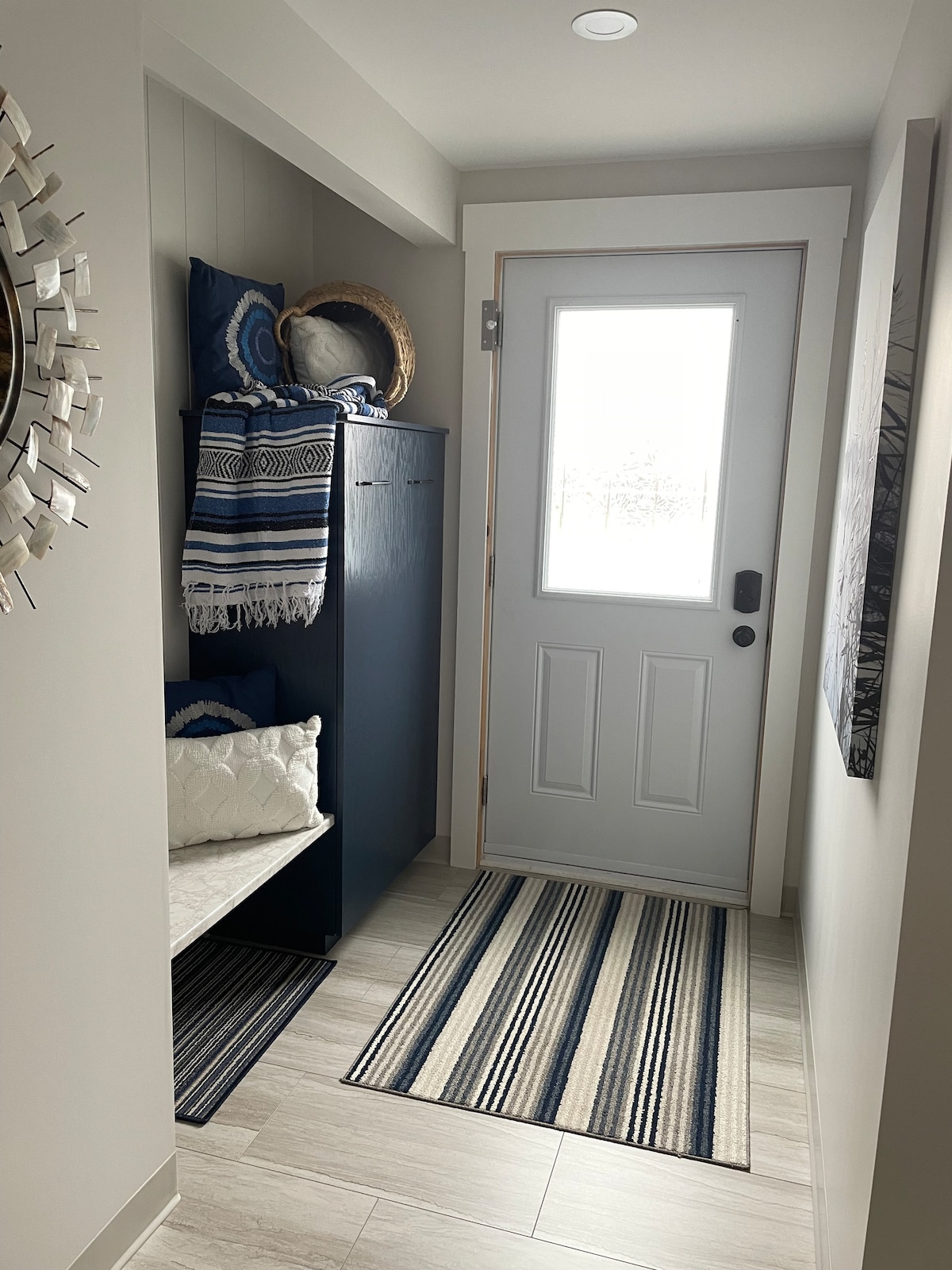 A well-organized entryway features a light-colored coat rack alongside a built-in bench. A striped rug extends across the tiled floor, complementing the blue hues of the decor. Natural light enters through a glass-paneled door, enhancing the welcoming feel of the space.