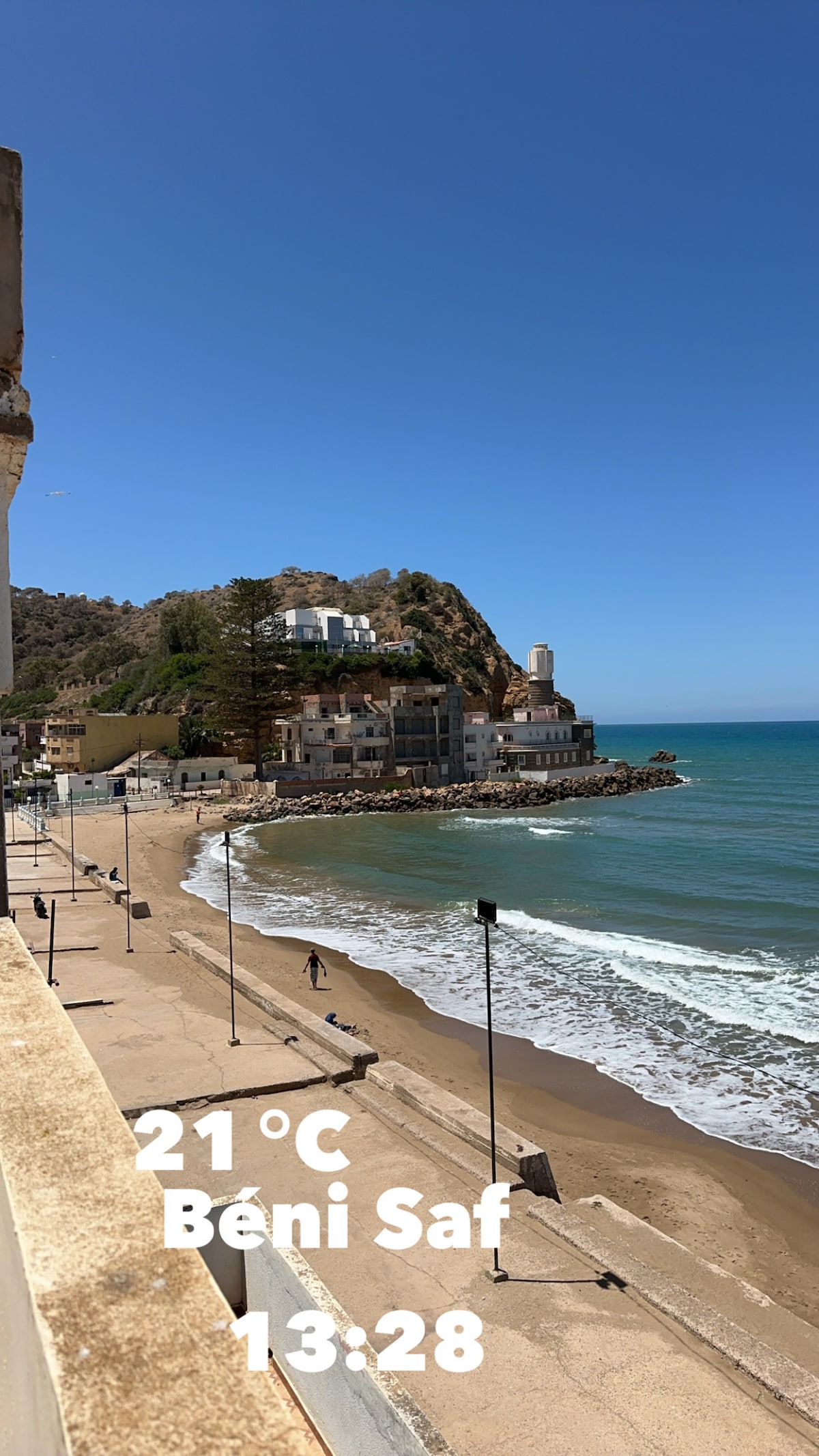 A coastal view captures a sandy beach adjacent to a calm sea, with gentle waves lapping at the shore. A hillside building is visible, along with a lighthouse in the background. The sky is clear, showcasing a serene atmosphere.