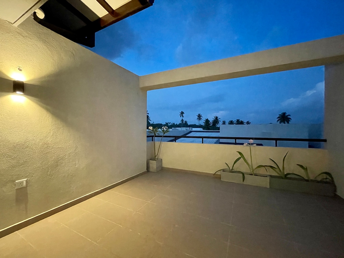 An open terrace is viewed under twilight, with soft lighting enhancing the clean, minimalist walls. A railing offers a glimpse of the landscape beyond, where gentle silhouettes of palm trees are visible against the evening sky. Potted plants add subtle greenery to the space.