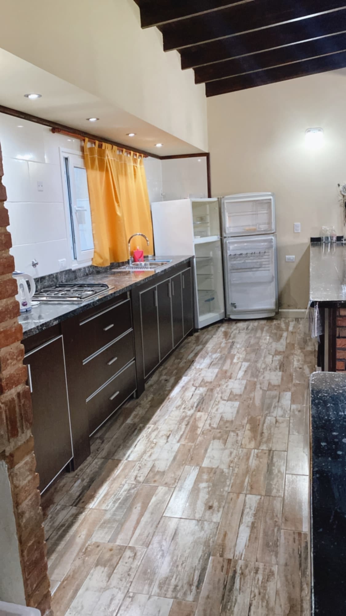 A modern kitchen showcases dark cabinetry against a light wall. A window with yellow curtains allows natural light, while a spacious countertop is equipped with a stove and sink. A refrigerator is visibly open, and tiled flooring adds a touch of elegance.