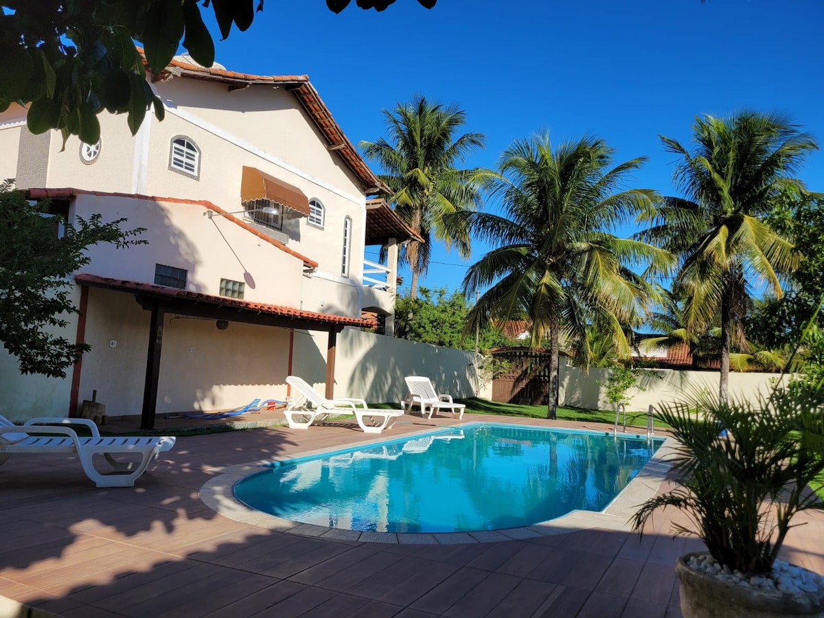 A spacious outdoor area features a clear swimming pool surrounded by palm trees and lounging chairs. The two-story house stands in the background, complemented by a blue sky overhead, adding to the inviting atmosphere of the space.