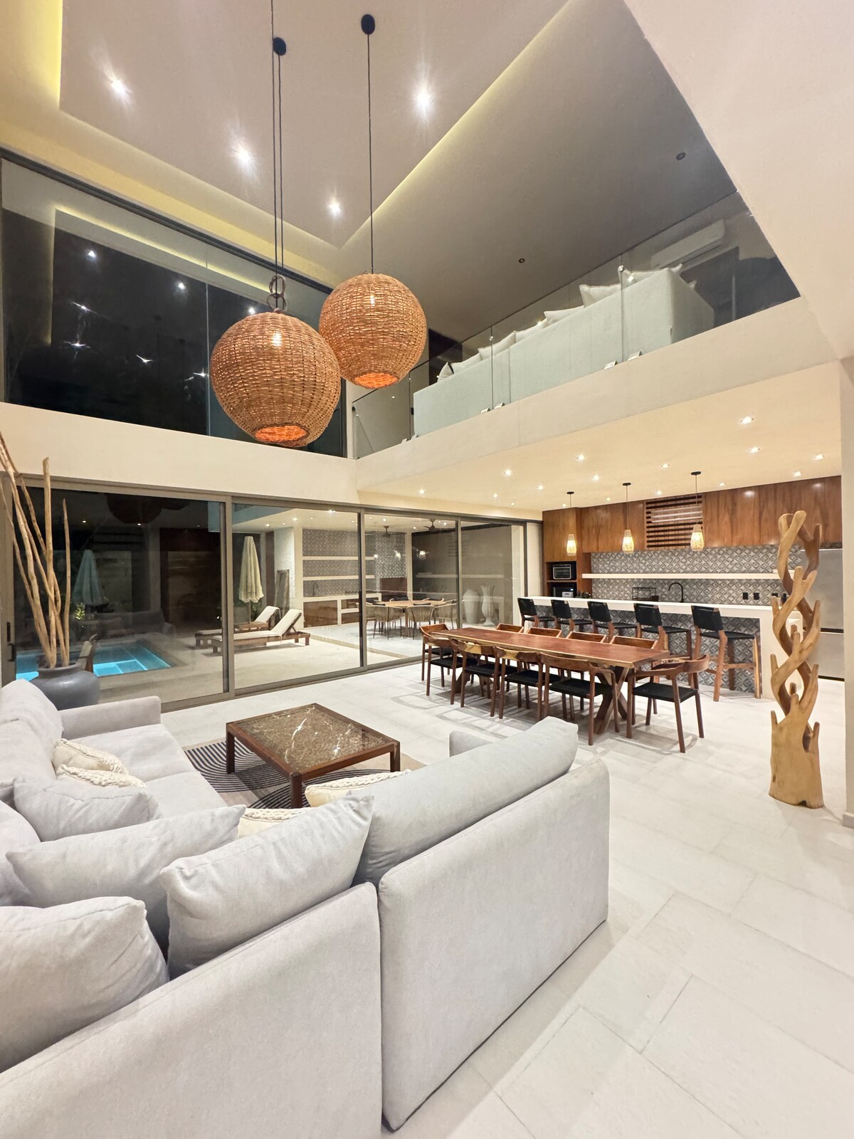 An open-concept living area displays a spacious light-toned sofa, paired with a wooden coffee table. Large glass doors provide a view of the outdoor pool area, while a dining table for several guests is situated nearby. Two pendant lights hang from the ceiling, highlighting the modern interior.