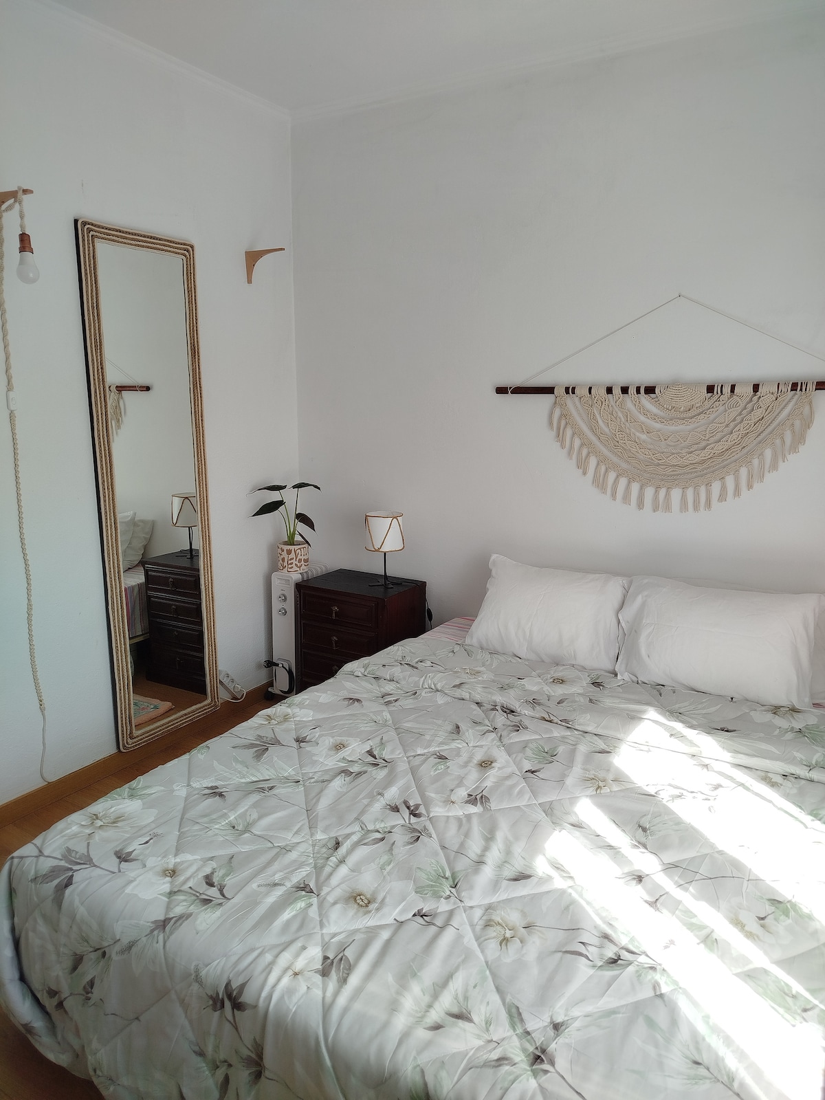 A bright bedroom is enhanced by natural light streaming through a window. A spacious bed with light bedding is positioned against a neutral wall. A standing mirror and a small nightstand are visible, along with a decorative wall hanging and a potted plant, creating a calming atmosphere.