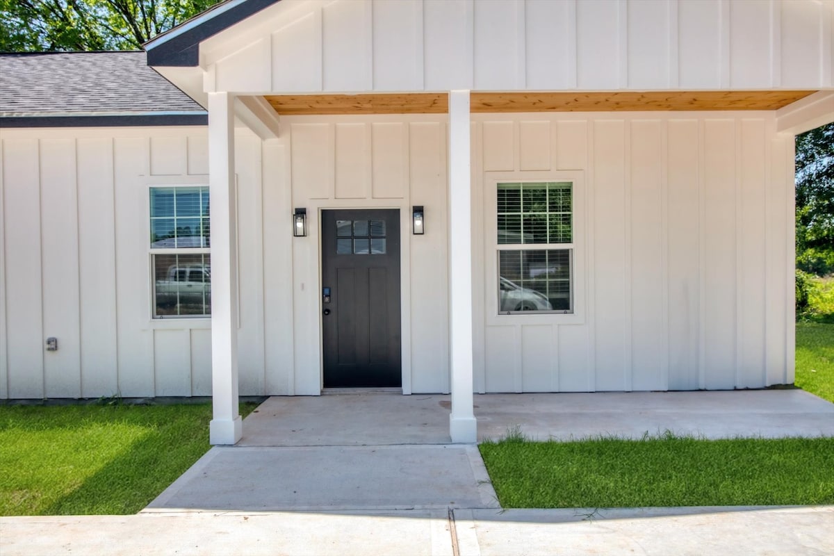 The front entrance of the unit features a dark door framed by two stylish light fixtures. A clean, concrete walkway leads to the entrance, bordered by neatly trimmed green grass. Large windows on either side allow natural light to illuminate the area.