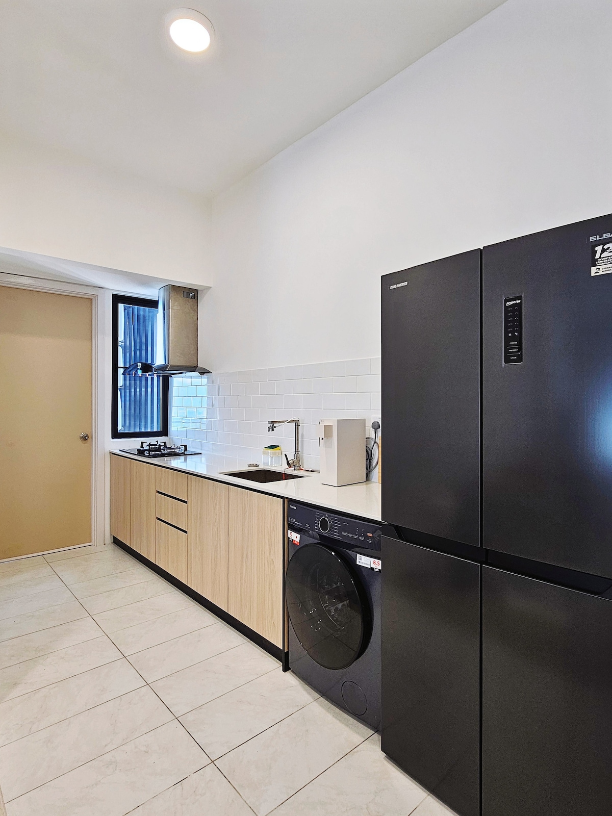 A modern kitchen features sleek cabinetry in light wood tones and white subway tile backsplash. Black appliances, including a refrigerator and washing machine, complement the neutral color scheme. Natural light enters through a small window, creating a bright and functional cooking space.