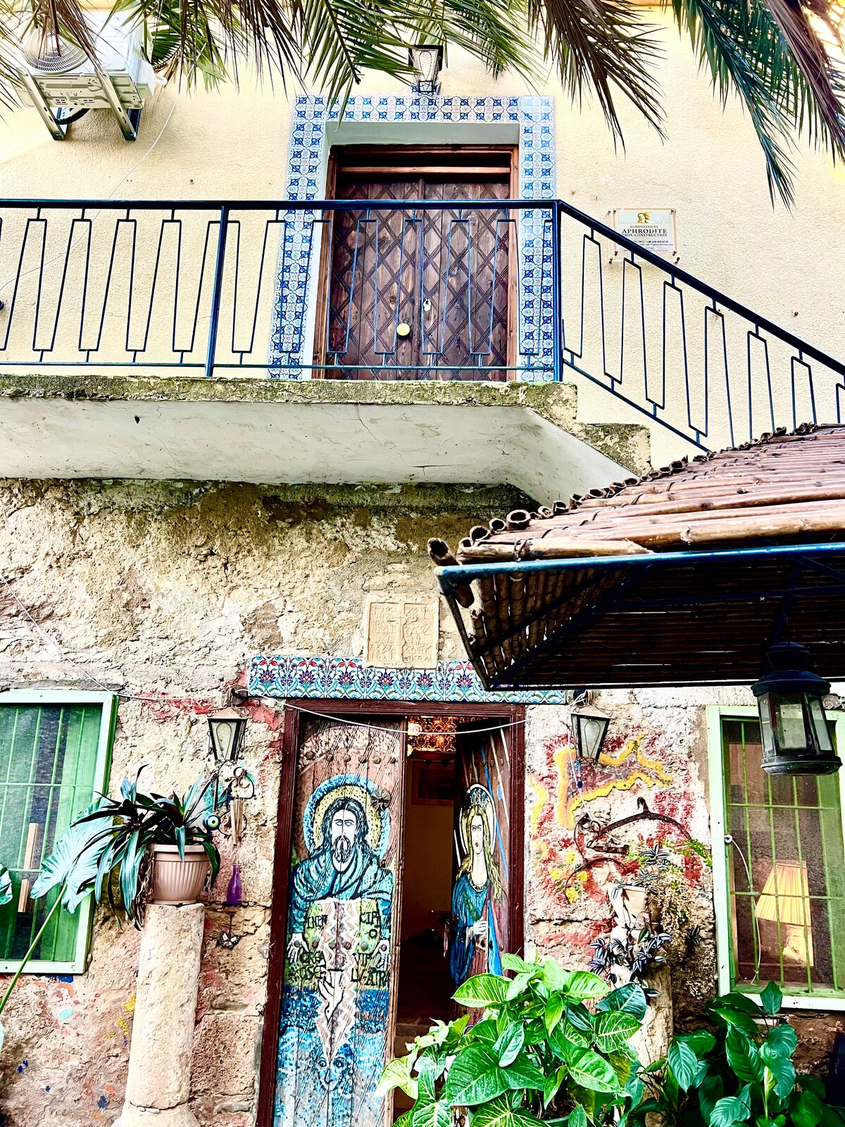 An entrance showcases a decorated door framed by colorful murals depicting figures. The textured wall features lush green plants and potted flora, while lanterns hang at either side, enhancing the welcoming appearance. A balcony above is bordered by a wrought iron railing.