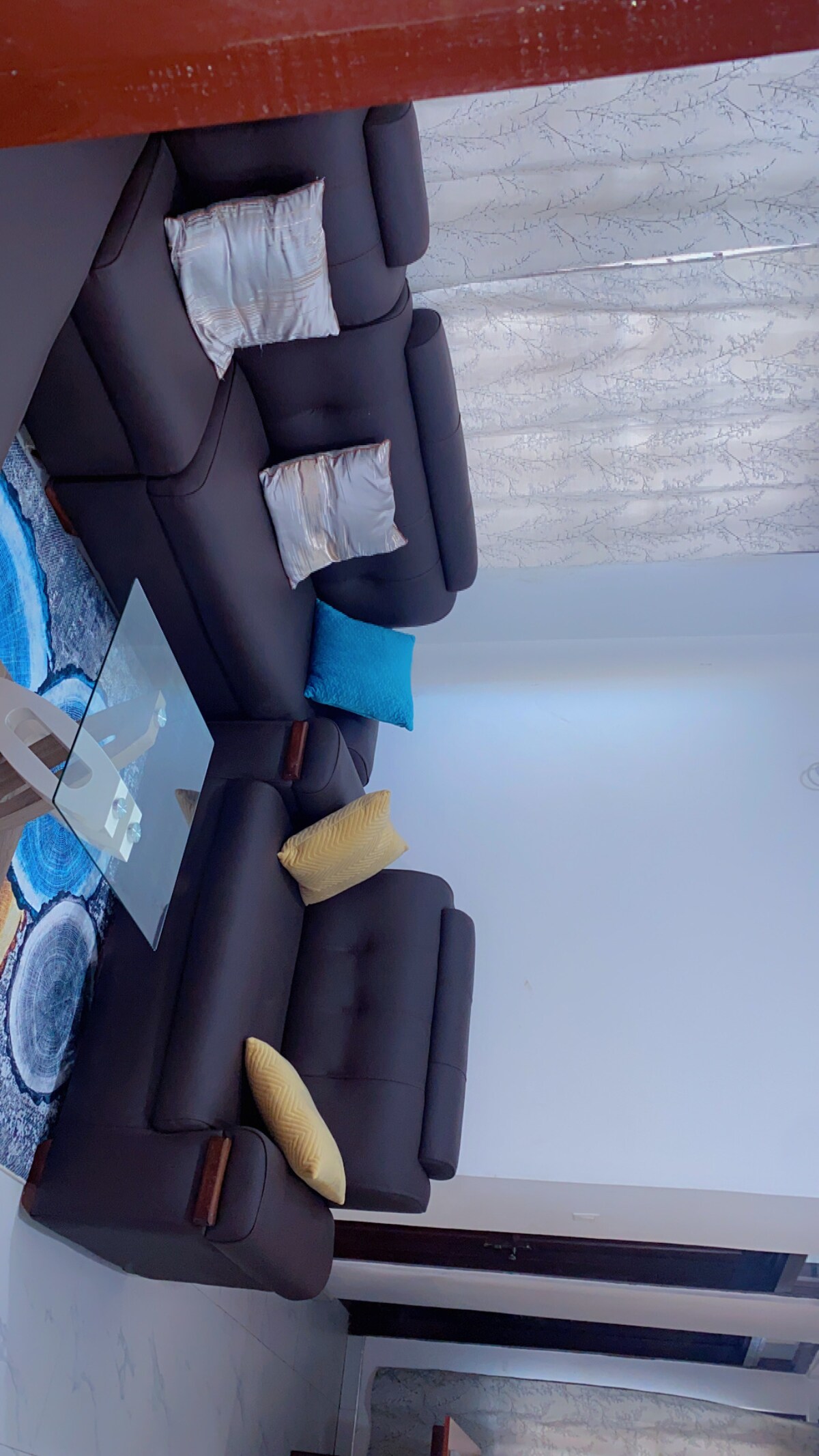 A comfortable seating area features a dark sectional sofa adorned with decorative pillows in shades of blue and yellow. A glass coffee table is centrally placed on a patterned area rug, with soft natural light filtering through sheer curtains.