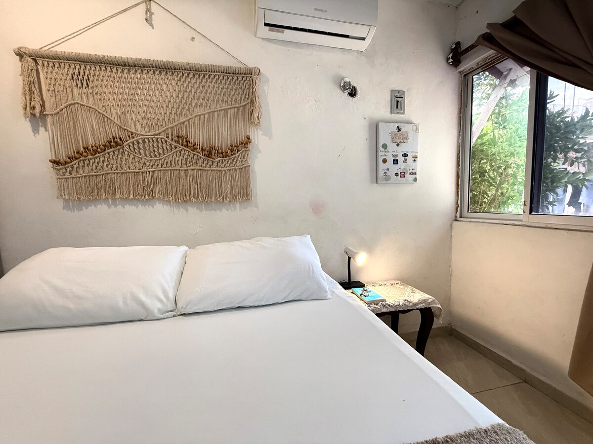 A simple and bright sleeping area features a spacious bed adorned with two white pillows. A macramé wall hanging adds texture to the light-colored wall. A small bedside table with a lamp sits next to the bed, and natural light enters through the window, providing a welcoming atmosphere.