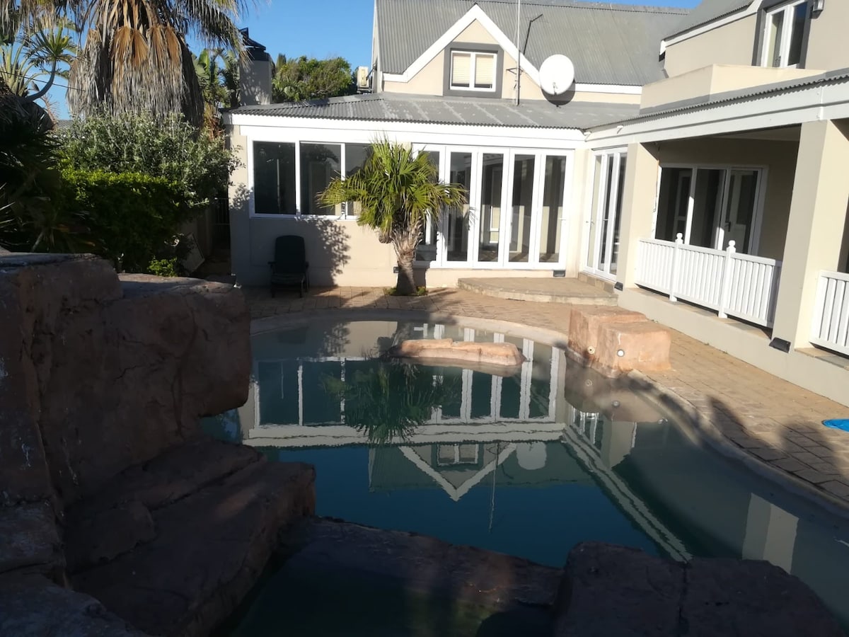 A private pool area is showcased, featuring rock formations and a serene, reflective water surface. Surrounding landscaping includes palm trees and a covered patio, while the modern home is visible in the background with large glass windows that bring in natural light.