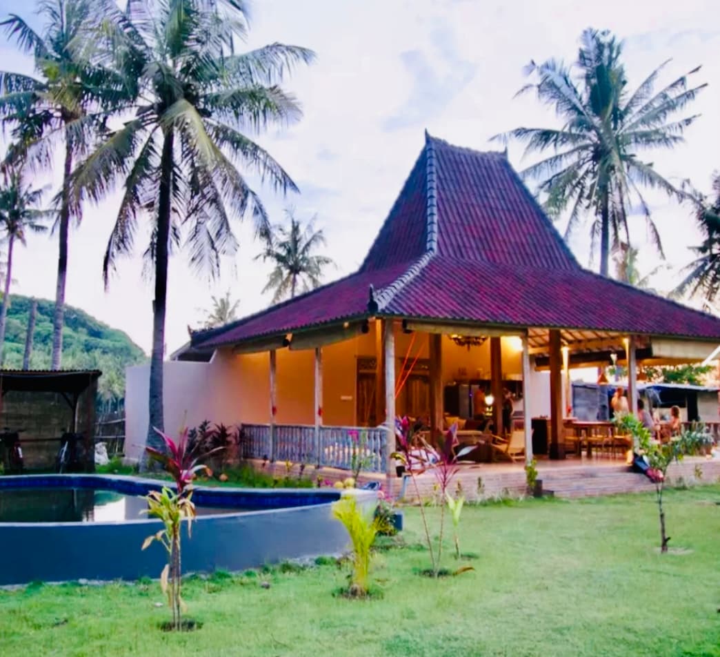 A traditional Javanese Joglo villa with a distinct roofline is surrounded by lush green grass and tropical plants. Coconut palms sway nearby, while an inviting pool reflects the sky, enhancing the villa's outdoor living space. The open-air design allows for natural light to illuminate the interior.