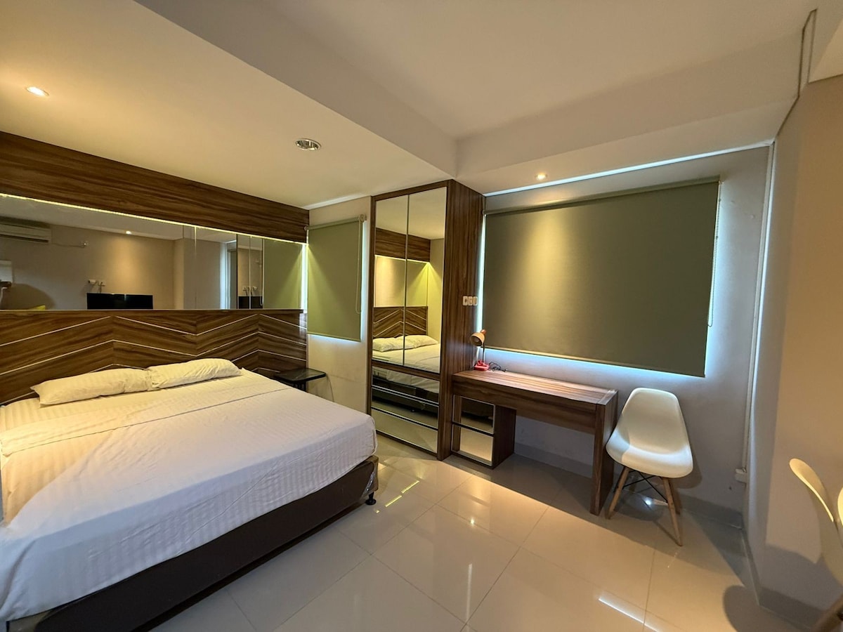 A modern sleeping area features a spacious bed dressed in white linens. A large mirror and a sleek desk are included, alongside a white chair. Natural light enters through partially closed window coverings, creating a bright and airy environment.
