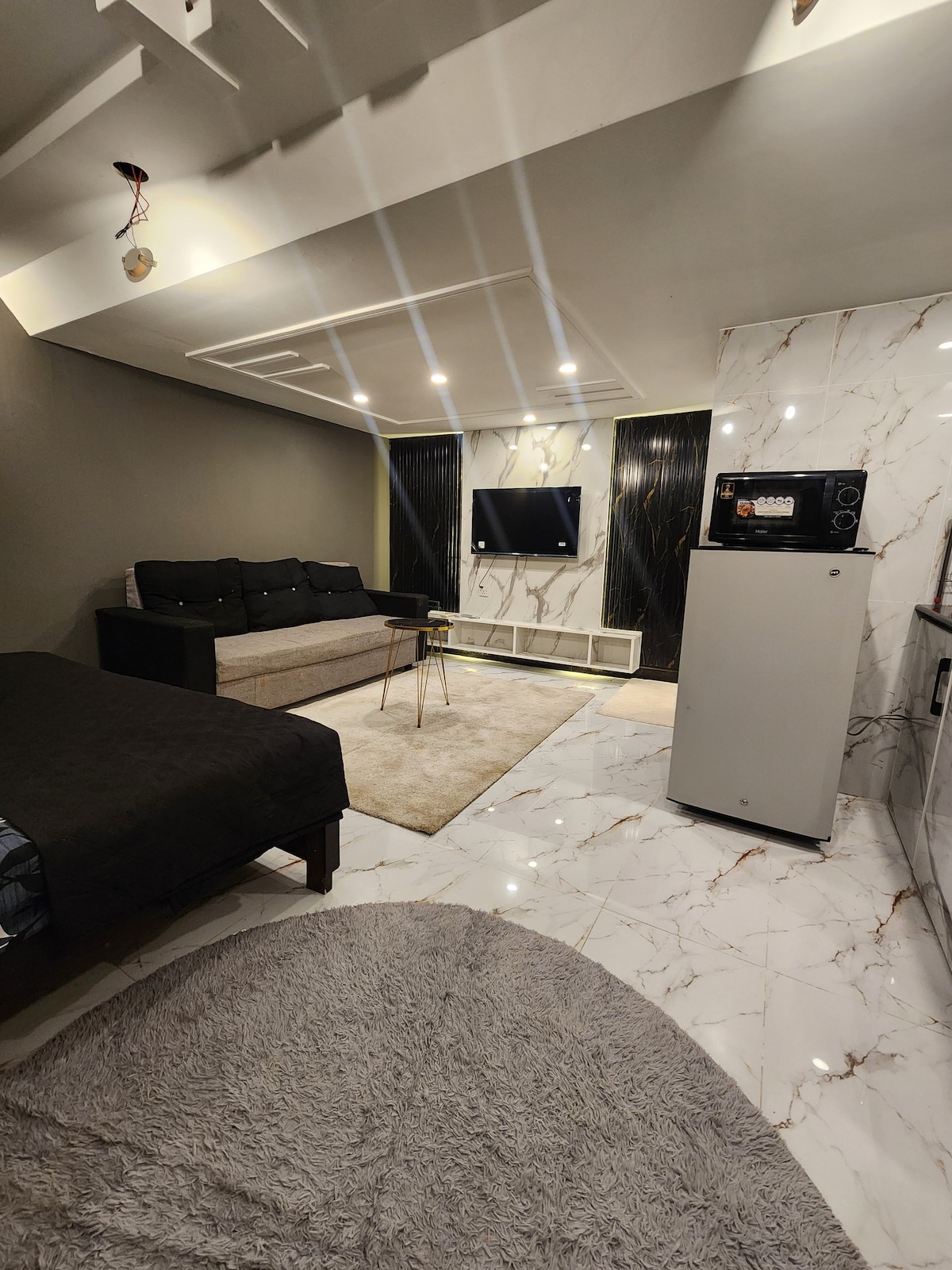 A modern studio apartment layout includes a cozy seating area with a dark sofa, a wall-mounted television, and a dining table with chairs. Light-colored marble flooring and soft area rugs contribute to the space's elegance, while ambient lighting creates a warm atmosphere.