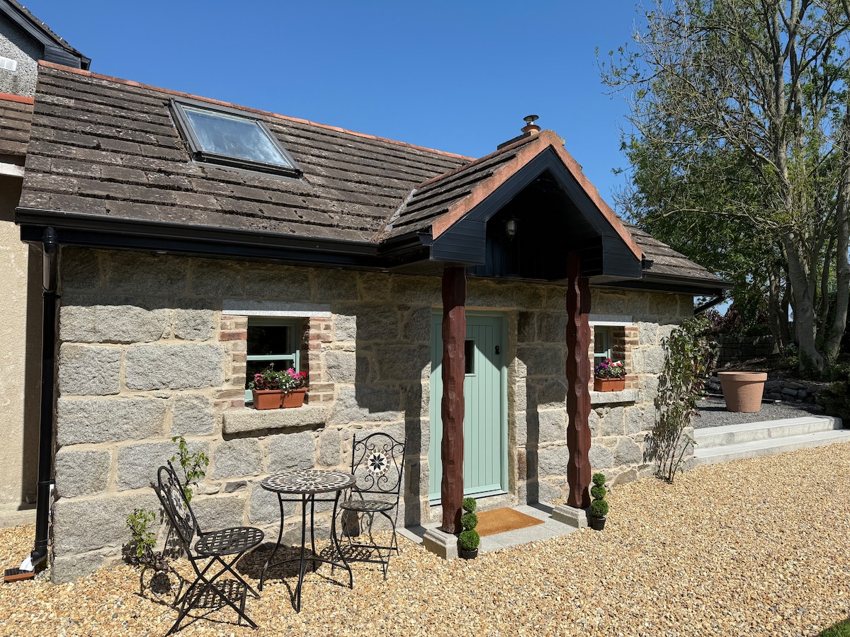 The exterior of the cottage features a combination of stone and wooden elements, presenting a welcoming entrance. Potted plants and flowers adorn the windows, while a small outdoor seating area with two chairs and a round table is positioned on a pebble-strewn pathway.