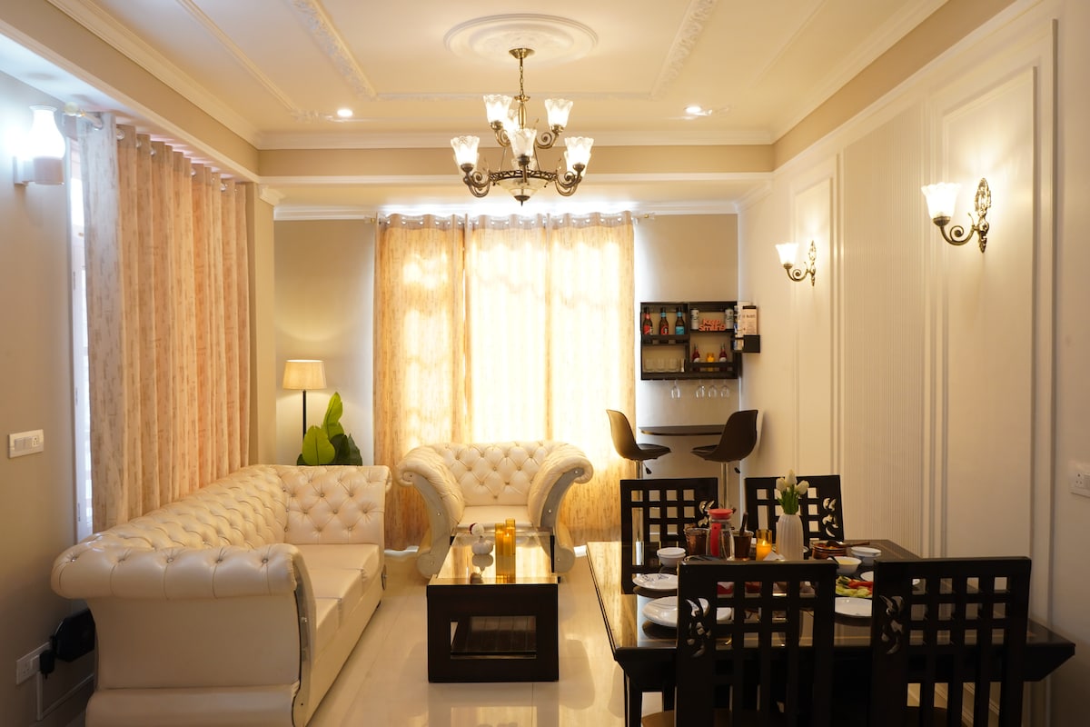The living and dining area features a bright, open layout with large windows allowing natural light to fill the space. A plush, white sofa is complemented by dark, wooden dining furniture. The chandelier and wall sconces provide warm lighting, creating a comfortable ambiance.