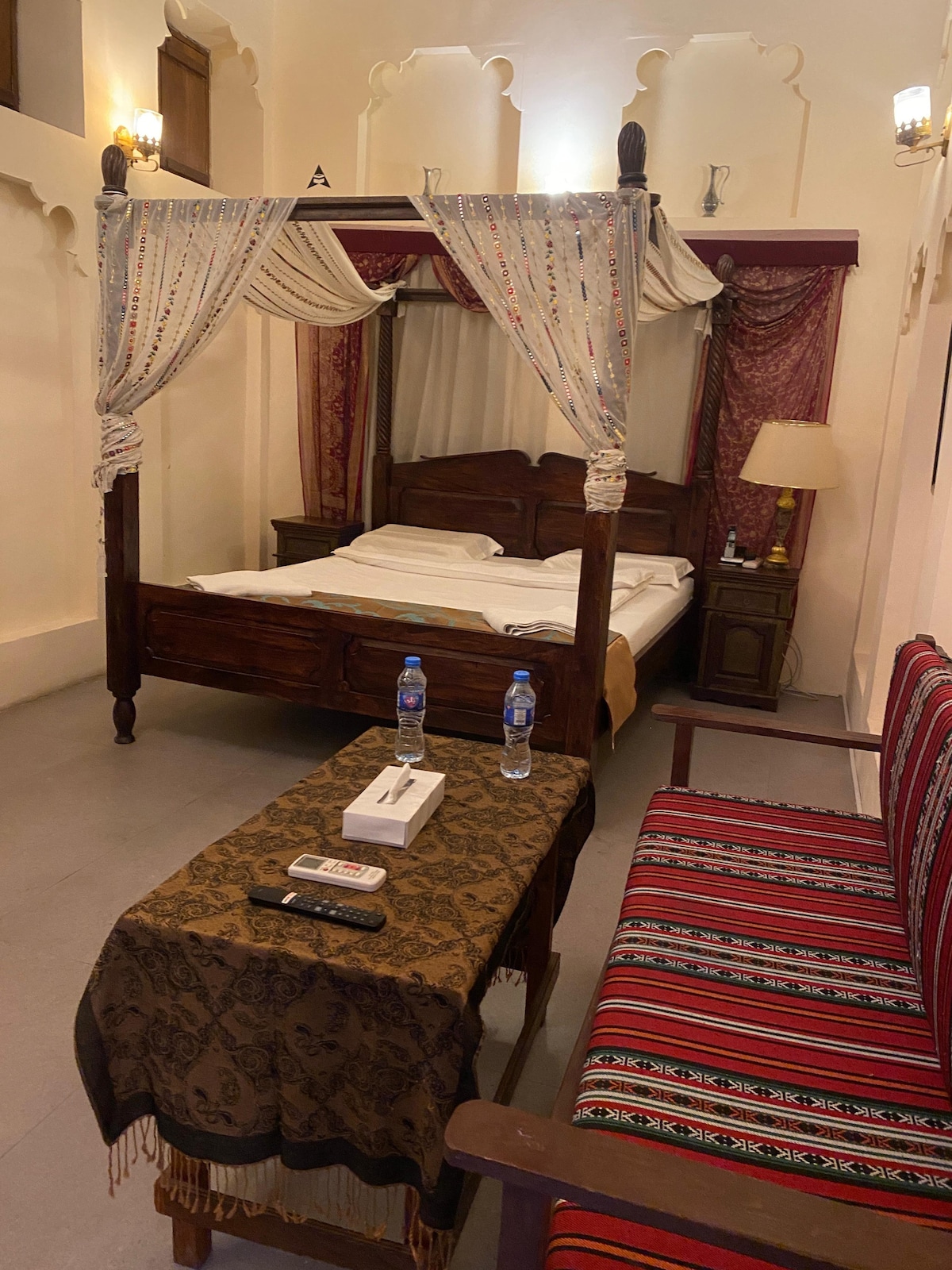 A wood-frame bed is draped with decorative fabric, complementing the intricate wall designs. A cozy seating area includes a striped sofa and a wooden table with refreshments. Ambient lighting is provided by lamps on either side of the bed, enhancing the space's welcoming feel.