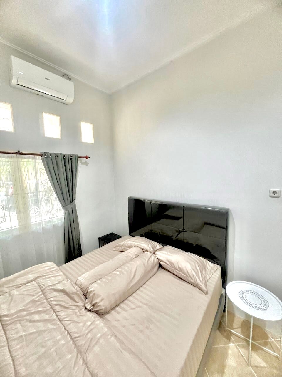 A modern bedroom features a sleek black headboard and a spacious bed with soft, neutral bedding. A small bedside table stands beside the bed, while large windows allow natural light to illuminate the room. Air conditioning is installed for comfort.