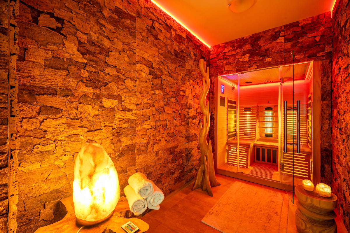 A wellness area features a rock wall illuminated by soft, warm lighting. A glass door leads to an infrared sauna visible in the background. Soft towels are neatly stacked next to a glowing stone, creating a serene ambiance for relaxation.