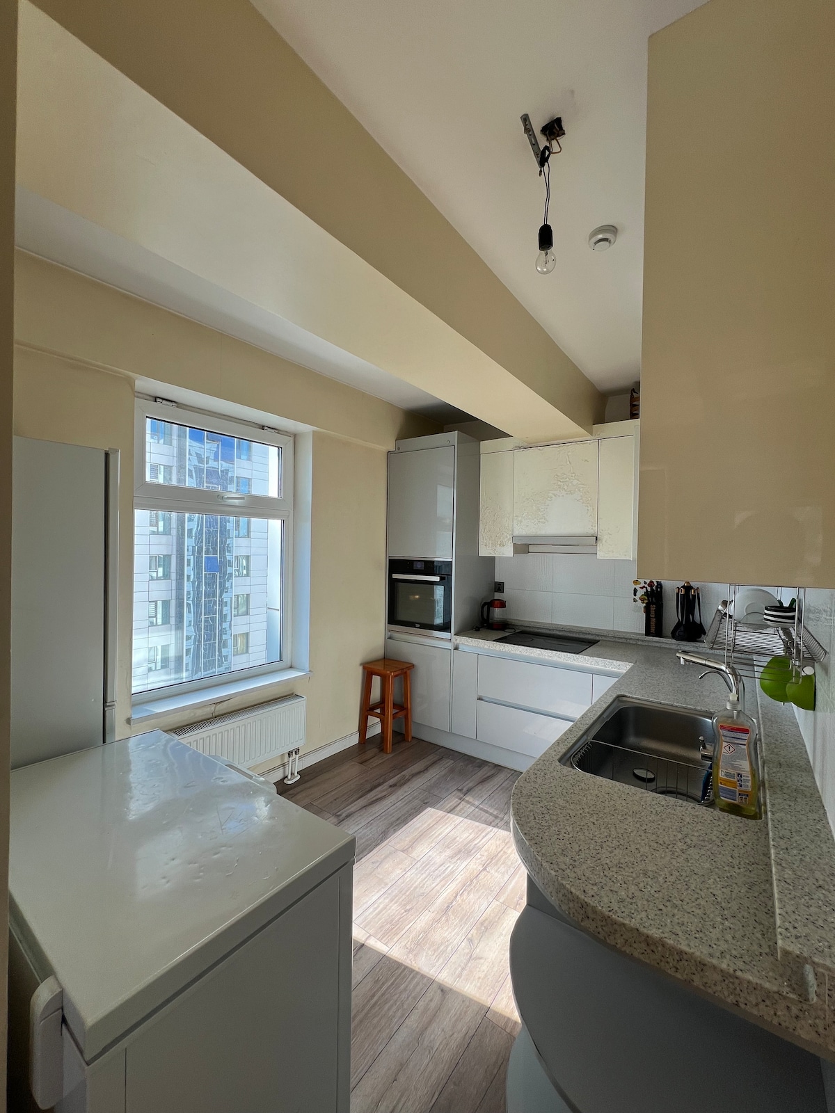 The kitchen is equipped with modern appliances, including a built-in oven, refrigerator, and washing machine. A window allows natural light to brighten the space, illuminating the sleek countertop and cabinetry. Ample storage is provided, and a minimalist design enhances the overall appearance.