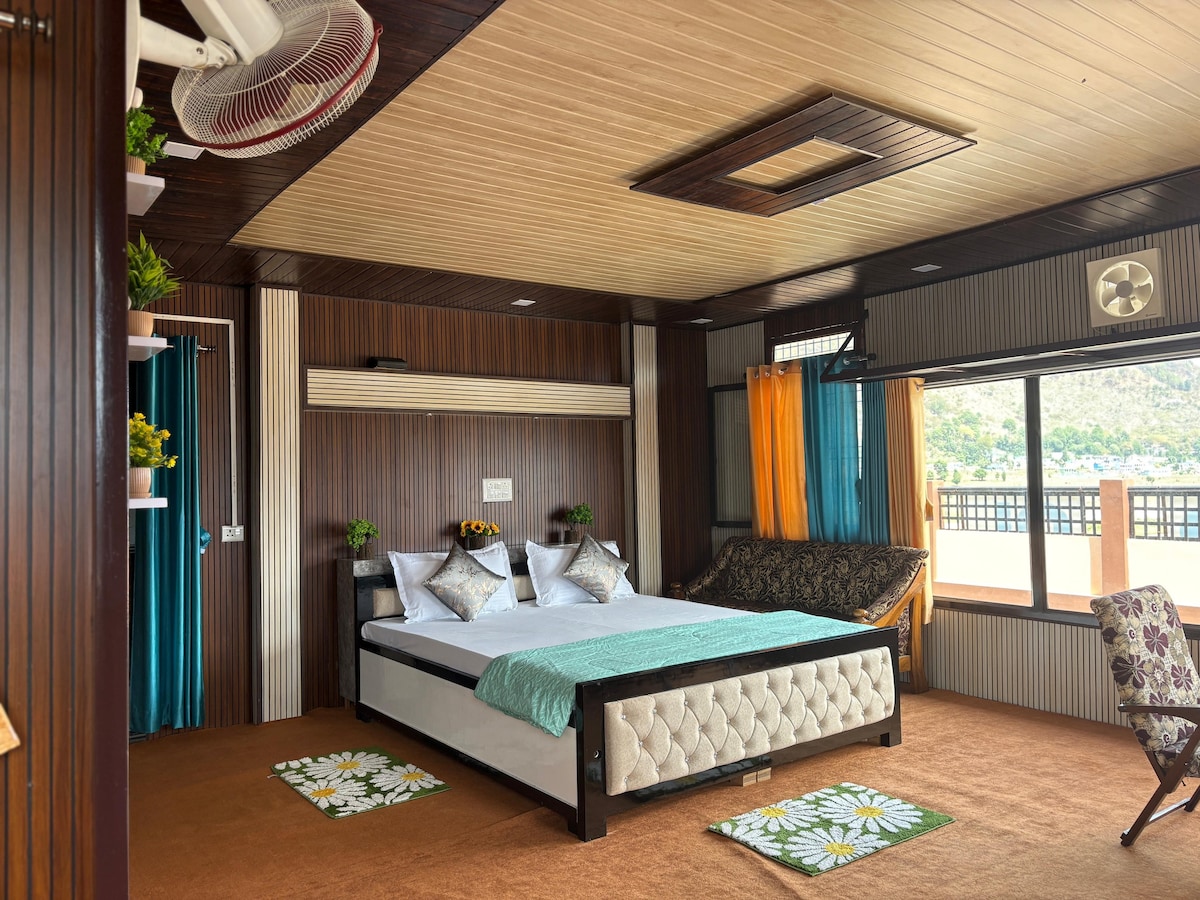 A spacious bedroom features a comfortable bed with textured bedding and decorative pillows. A seating area with a patterned sofa is present beside the large window, which offers views of surrounding greenery. Vibrant curtains frame the window, and decorative elements enhance the room's character.