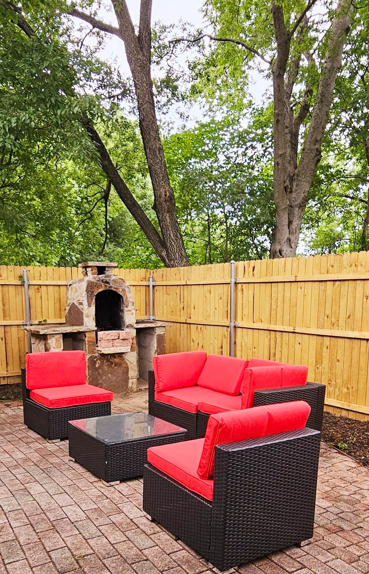 A cozy patio space features a seating arrangement with red cushions on black wicker chairs surrounding a glass-topped table. A stone outdoor fireplace stands nearby, while a wooden fence and greenery provide privacy and a natural setting.