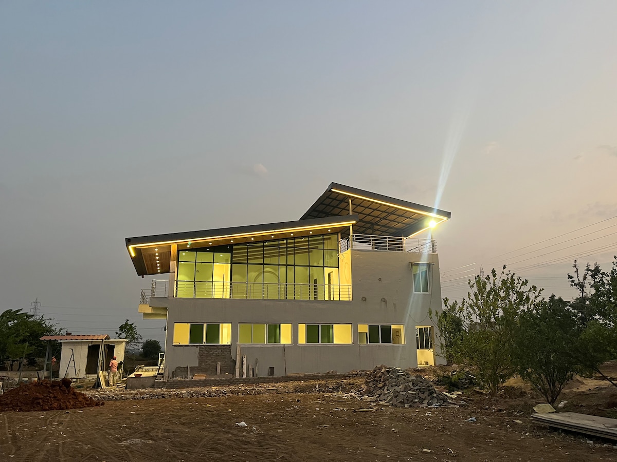 A modern two-story structure showcases large glass windows reflecting warm light. A distinctive sloped roof adds visual interest, while surrounding open land features a mix of cleared space and natural vegetation.