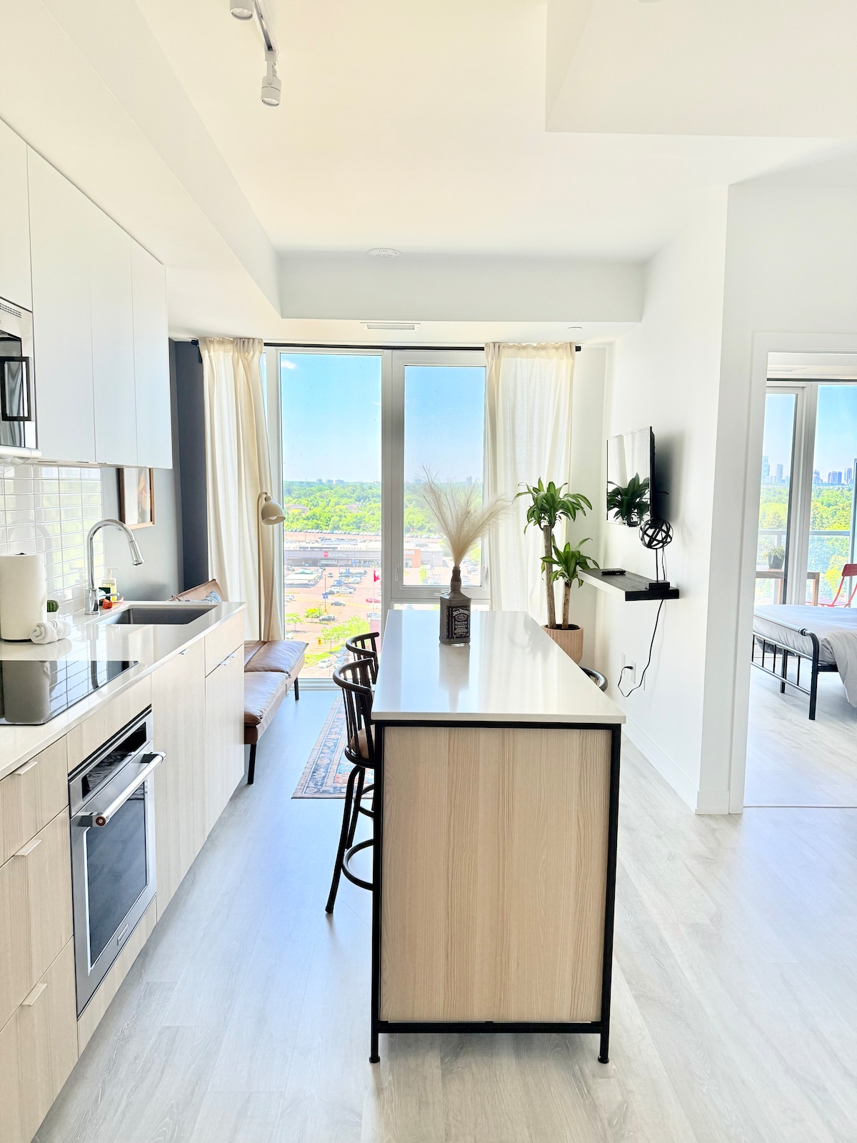 A modern kitchen is visible, featuring sleek cabinetry and stainless steel appliances. A spacious island with bar stools offers a casual dining area. Large windows provide natural light and showcase views of greenery and the city skyline. Decorative elements include a plant and a wall-mounted television.