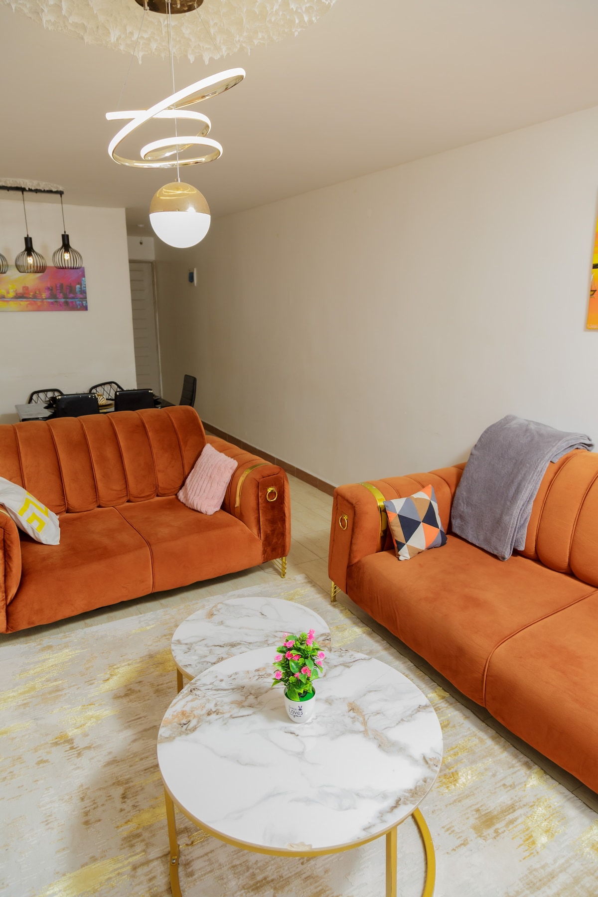 A comfortable seating area features two orange sofas adorned with decorative pillows. A marble coffee table sits in the center, complemented by a small vase of flowers. The space is softly illuminated by modern pendant lights, while muted wall art adds a splash of color.