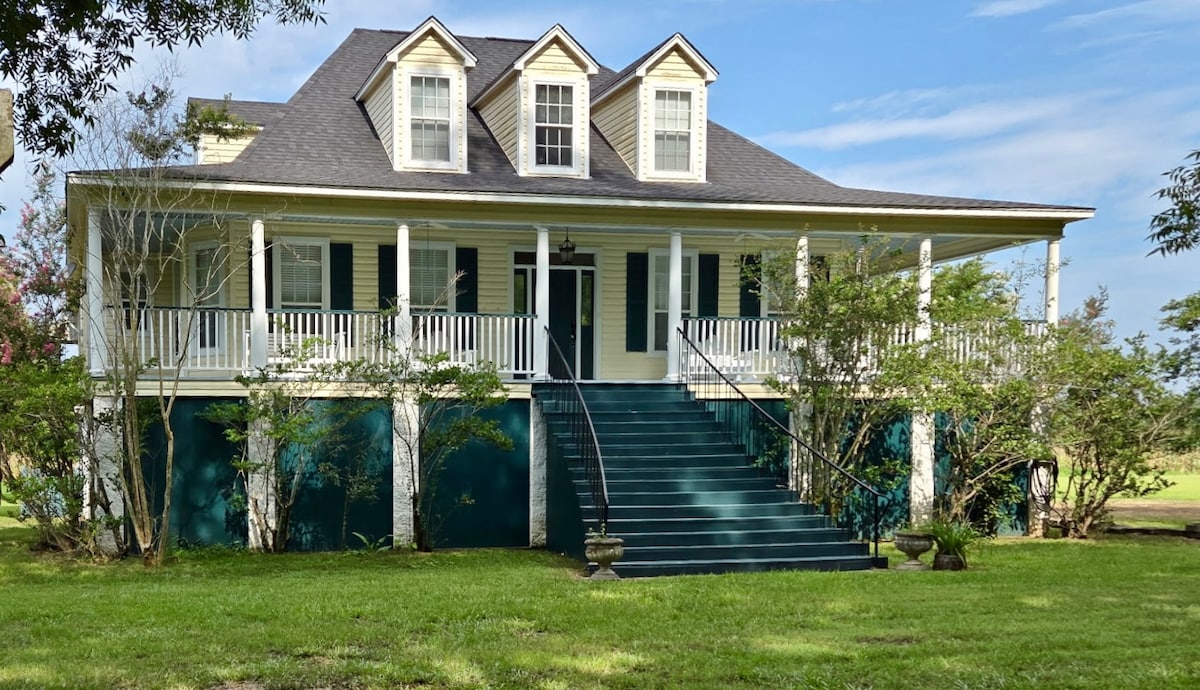 A farmhouse with a spacious wraparound porch is presented. Stairs lead up to the main entrance, which is flanked by multiple windows. The exterior is painted in soft hues, with greenery surrounding the property, hinting at the rural landscape that adds to its charm.